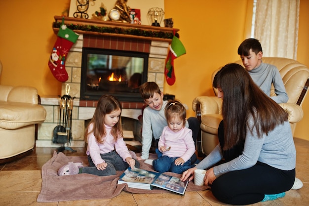 Happy young large family by a fireplace in warm living room on winter day Mother with four kids at home read book