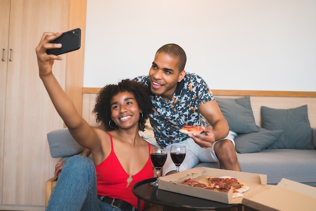 Happy young latin couple taking a selfie with smartphone while sharing a food together at their home. Lifestyle concept.