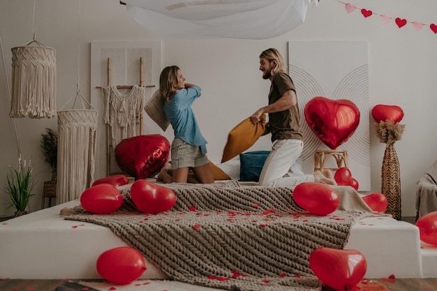 Happy young loving couple having a pillow fight in bed surrounded with red heart shape balloons