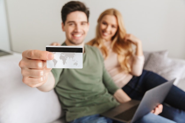 Happy young loving couple sitting on sofa holding debit card.