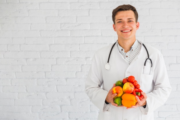 Happy young male doctor standing against the wall holding fresh fruits and vegetable