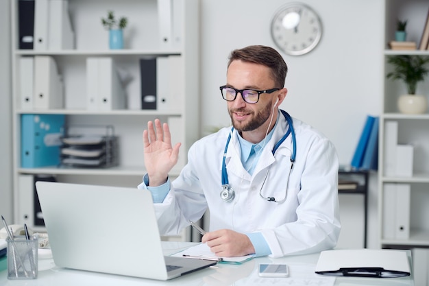 Happy young male doctor in whitecoat and eyeglasses greeting online patient by waving hand while sitting in front of laptop in medical office