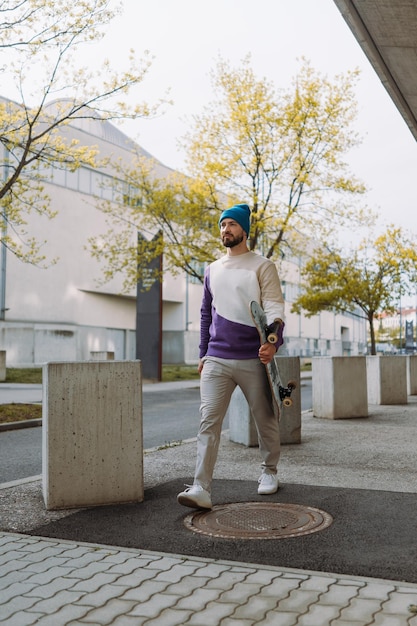 Happy young male skater in casual outfit carrying skateboard while walking along asphalt road vertical photo High quality photo