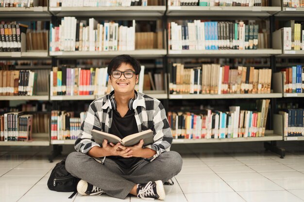 Happy young male student wearing eyeglasses and reading book while crossed legs sitting in library