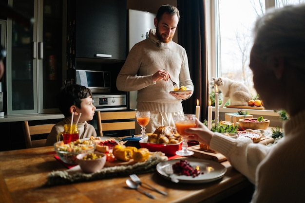 Happy young man arranging food on plates to wife and son standing at festive christmas table during