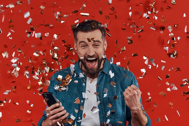 Happy young man in casual clothing holding smart phone and cheering while standing  with confetti flying around