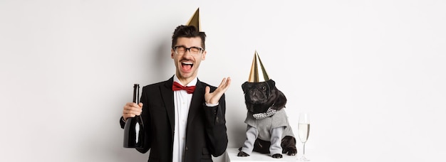 Happy young man celebrating holiday with cute dog holding champagne and smiling pug and owner wearin