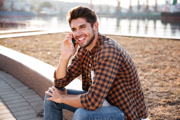 Happy young man in checkered shirt sitting and talking on cell phone in port