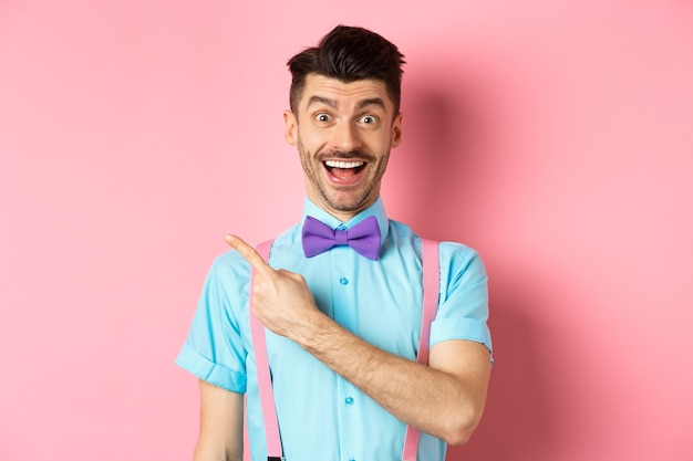 Happy young man checking out promo offer, smiling amazed and pointing finger left at copy space, standing near logo on pink background.