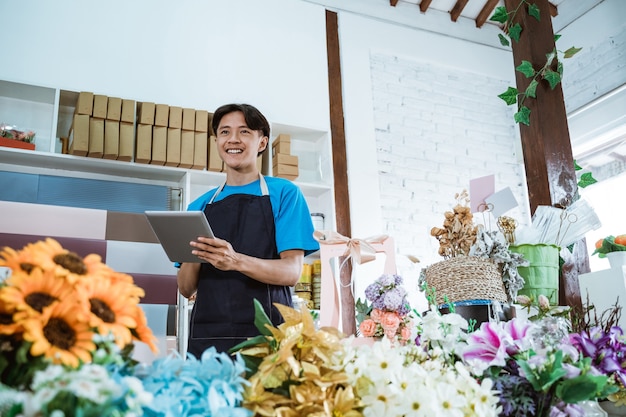 Happy young man entrepreneur working in flower shop wearing apron smiling while holding tablet