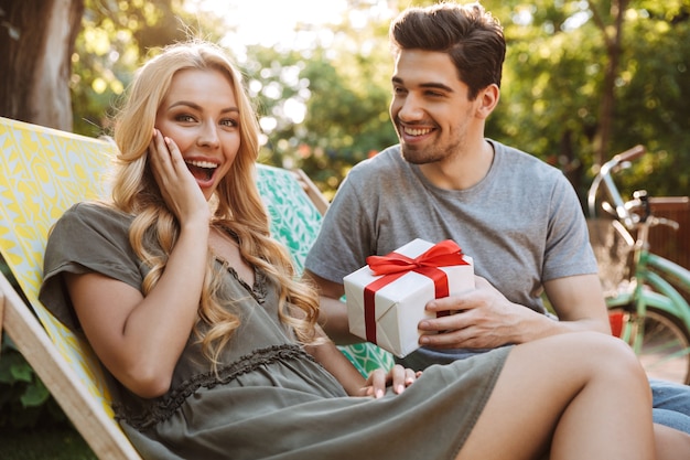 Happy Young man giving a gift box to his surprised happy woman while their sitting on sun loungers outdoors