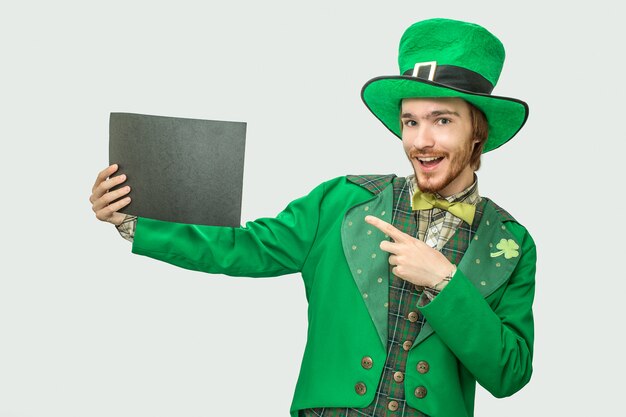 Happy young man in green suit holding black piece of paper. He wears saint Patrick's suit. Isolated on grey .