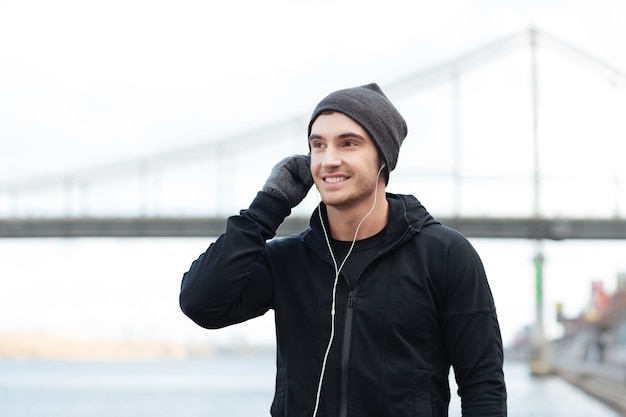 Happy young man in hat and gloves listening to music with earphones outdoors