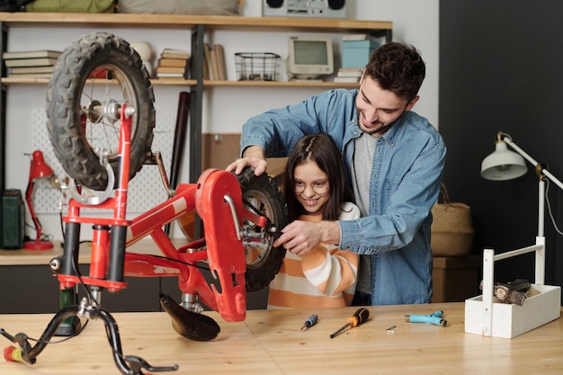 Happy young man and his daughter using wrench to fix part of bicycle wheel