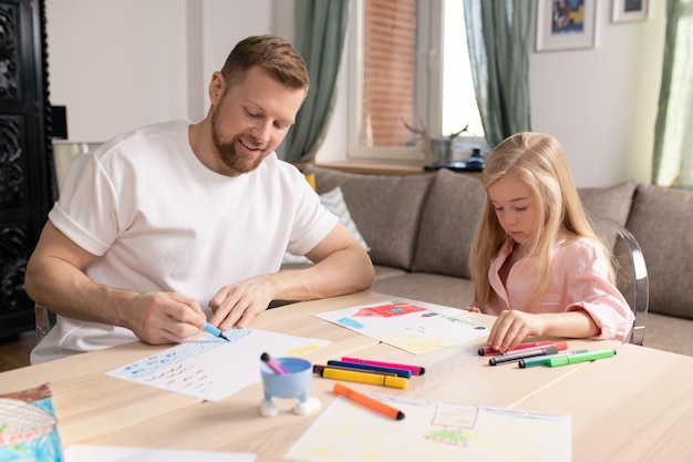 Happy young man and his little cute daughter sitting by wooden table in living-room and drawing with highlighters on paper