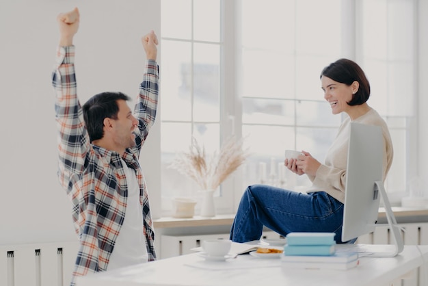 Happy young man rejoices successfully finished work, raises arms, dressed in checkered shirt, being glad, shares his success with wife who sits on desktop with coffee cup, collaborate together