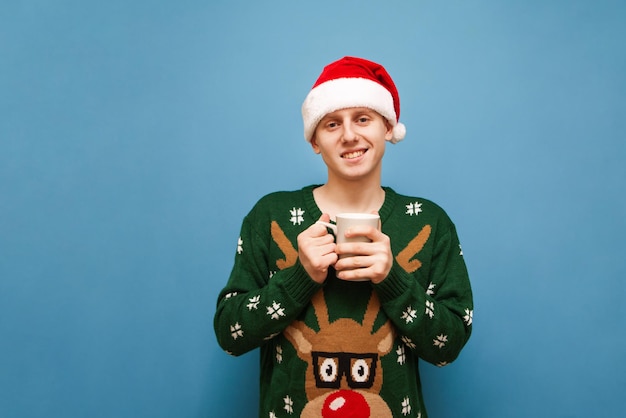 Happy young man in Santa hat and Christmas sweater with deer stands on blue background