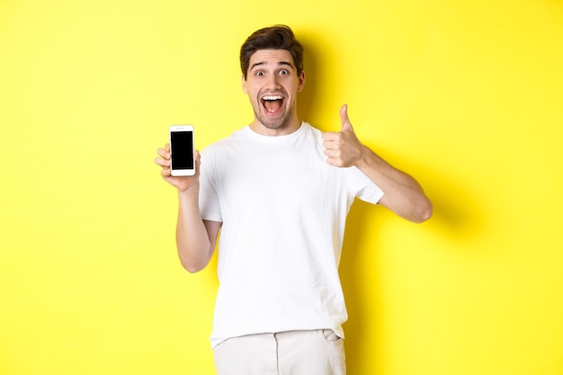 Happy young man showing thumb up and mobile phone screen, recommending application or internet website, standing over yellow wall