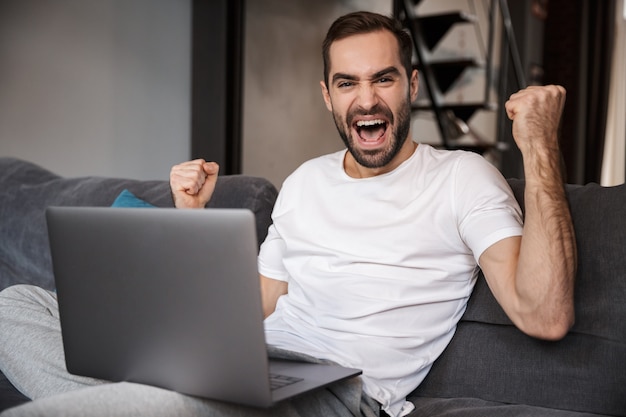 Happy young man sitting on a couch, using laptop computer, celebrating