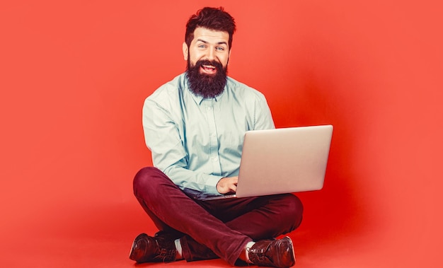 Happy young man sitting on the floor with and using laptop computer on red background Holding laptop computer