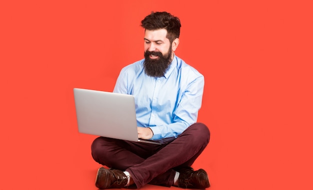 Happy young man sitting on the floor with and using laptop computer on red background.