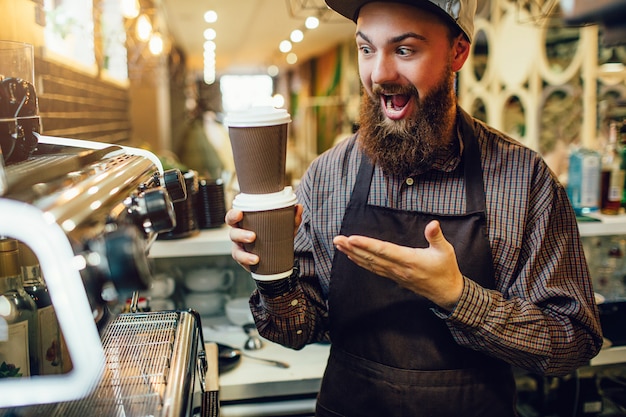 Happy young man stands at coffee machine and holds cups of hot drink