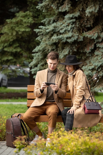 Happy young man in stylish warmwear and his girlfriend sitting on bench by coniferous tree in park and watching online video in smartphone