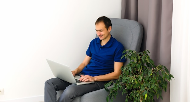 Happy young man in t-shirt sitting on sofa at home, working on laptop computer, smiling.