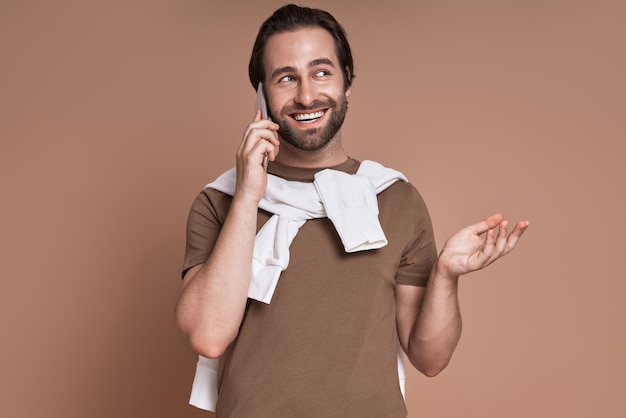 Happy young man talking on mobile phone and gesturing while standing against brown background