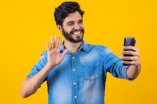 Happy young man video calling talking online with mobile phone saying hello to smartphone camera and waving hand friendly standing over yellow background