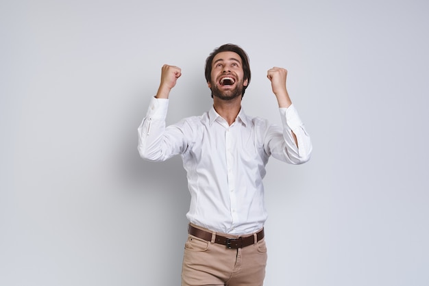 Happy young man in white shirt gesturing and looking up while standing against gray background