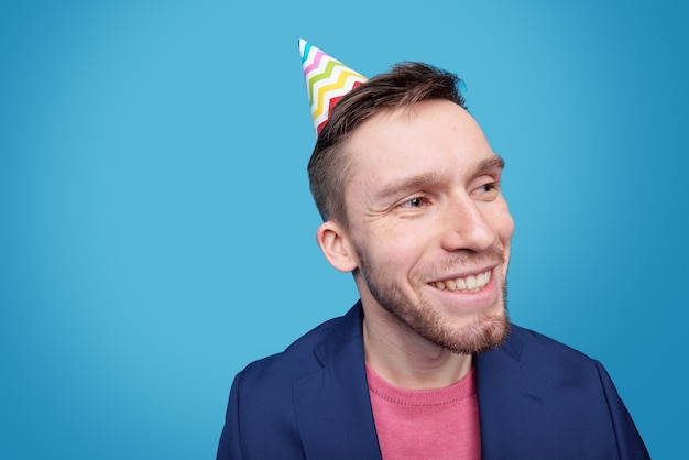 Happy young man with birthday cap on head looking aside with toothy smile while enjoying festive event