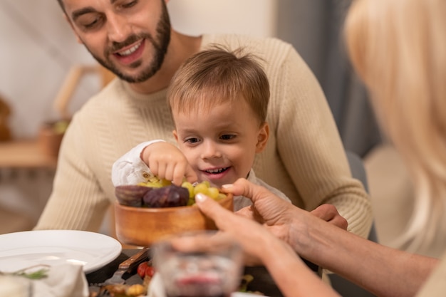 Happy young man with cute little son on his knees looking at him taking fresh grape from wooden bowl held by mature woman during dinner