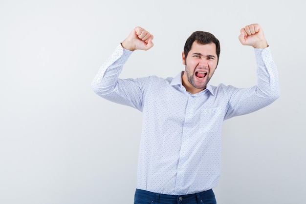 Happy young man with open mouth screaming and holding his fists above on white background