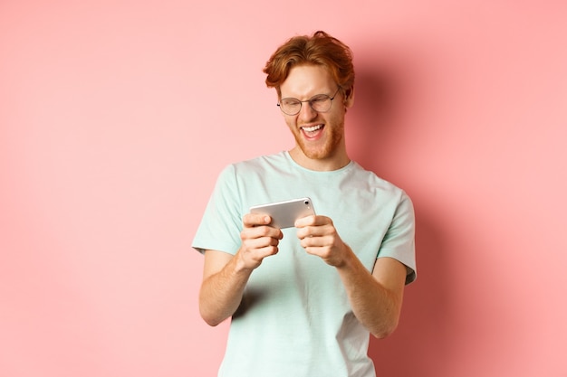 Happy young man with red messy haircut, wearing glasses, playing video game on smartphone and having fun, looking at mobile screen, standing over pink background