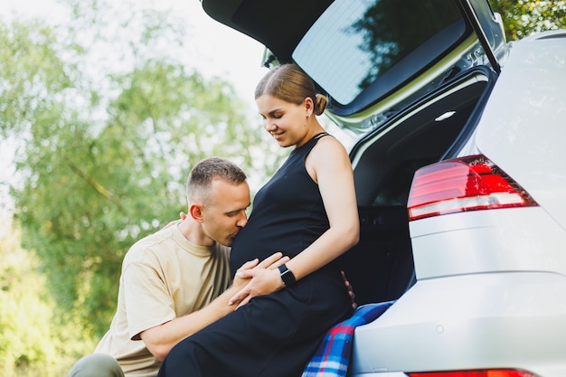 A happy young married couple expecting a child are sitting in the trunk of a car in nature A man and a woman hug a pregnant belly while sitting on a green lawn