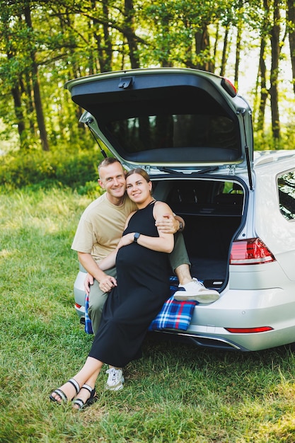 A happy young married couple expecting a child are sitting in the trunk of a car in nature A man and a woman hug a pregnant belly while sitting on a green lawn