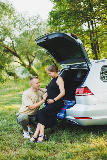 A happy young married couple expecting a child are sitting in the trunk of a car in nature A man and a woman hug a pregnant belly while sitting on a green lawn