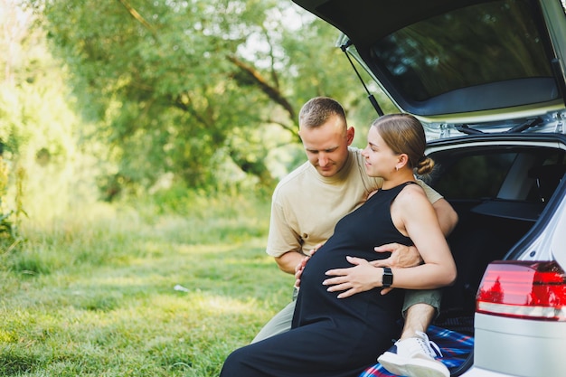 A happy young married couple expecting a child are sitting in the trunk of a car in nature A man and a woman hug a pregnant belly while sitting on a green lawn