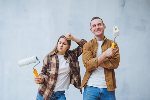 Happy young married couple in love in shirts doing renovations renovating walls painting with white paint roller preparing to move into new house selective focus