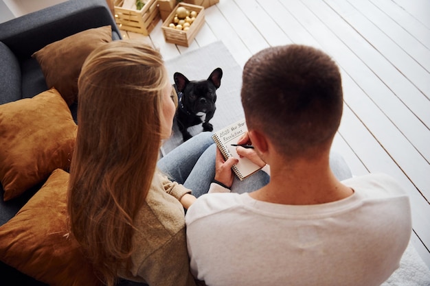 Happy young modern couple sitting on the sofa and writing wishes on notepad at home with their cute dog.