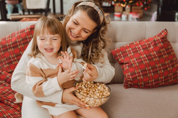 Photo happy young mom and baby girl laugh while sitting on the couch and eat popcorn. family watching tv and eating popcorn.