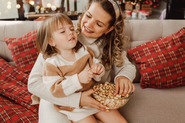 Photo happy young mom and baby girl laugh while sitting on the couch and eat popcorn. family watching tv and eating popcorn.