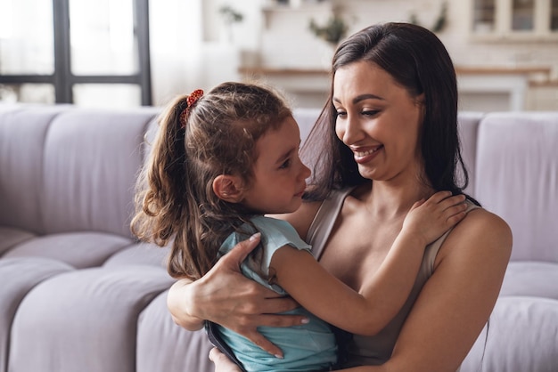 Happy young mother and her little daughter spend time together and have fun sitting hugging on the couch in the living room at home