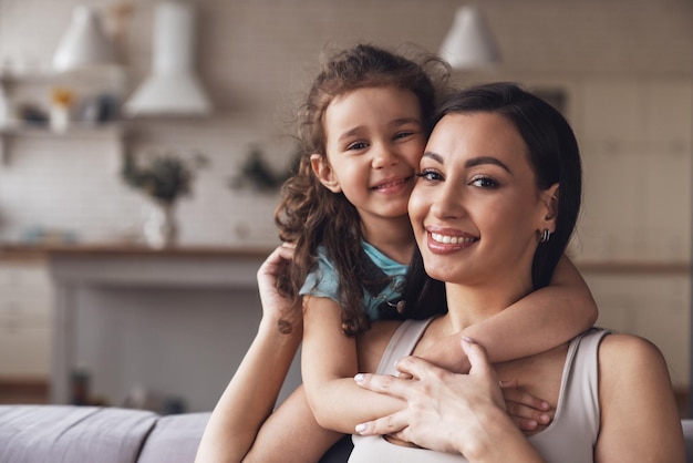 Happy young mother and her little daughter spend time together and have fun sitting hugging on the couch in the living room at home