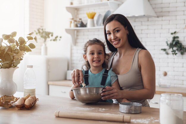 Happy young mother and little baby girl prepare dough and have fun while making cookies in the kitchen at home