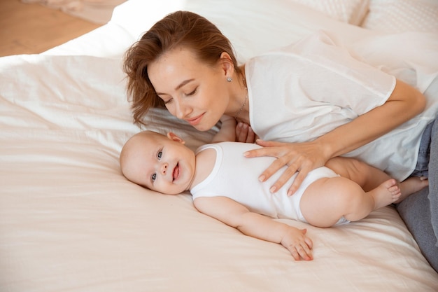 Happy young mother with baby in white room mom are lying kissing baby at home on bed in bedroom