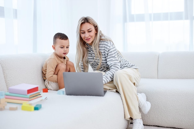 Happy young mother with little kid sit on sofa in living room have fun using modern laptop together ...