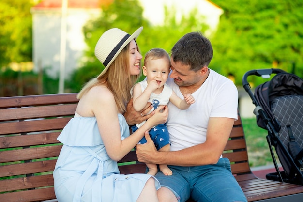 Happy young parents mom and dad with their baby son walk in the park in the summer on a bench and have fun and smile