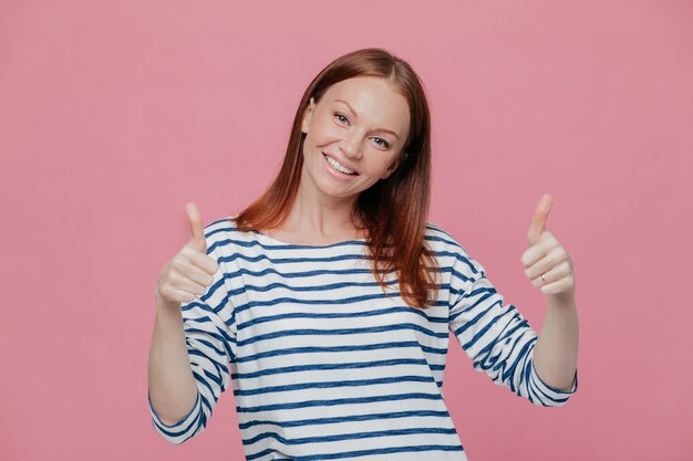 Photo happy young pretty european lady give double thumb up likes somebody idea or gives approval dressed in striped outfit isolated over pink background people body language gesturig concept